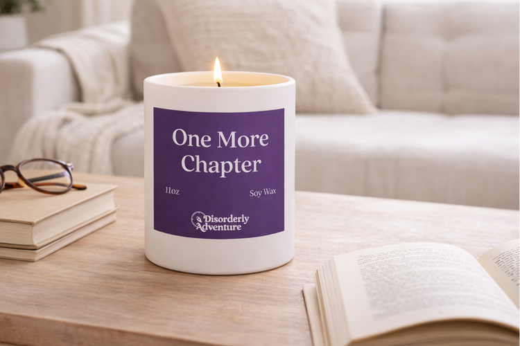 Candle labeled 'One More Chapter' on a table with books and glasses in a cozy living room.